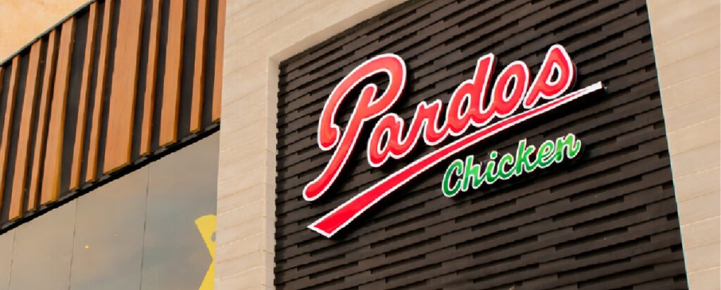 Fachada de Pardos Chicken con revestimiento oscuro, detalles en madera y letrero iluminado.